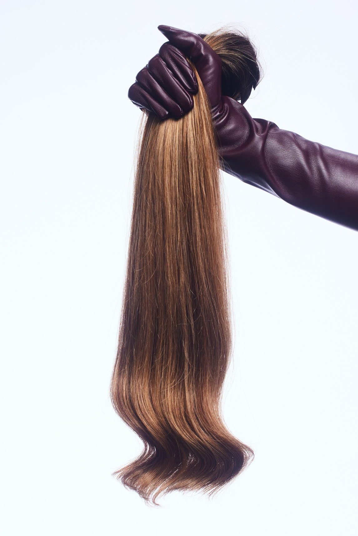 A long, straight hair extension piece with warm brown to light brown ombré tones, held up by a hand wearing a dark burgundy leather glove against a solid white background.