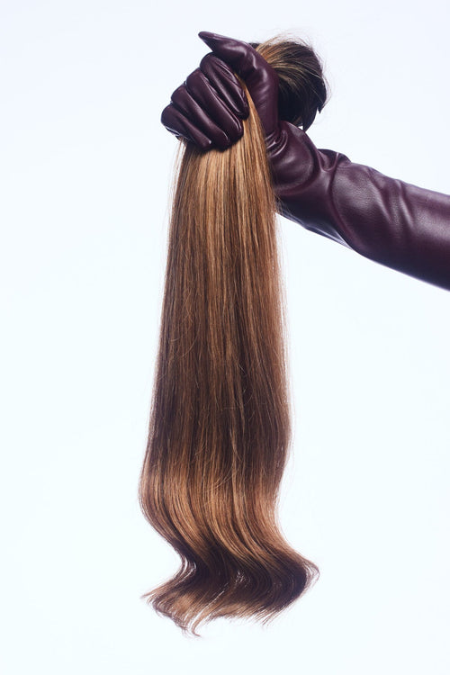 A long, straight hair extension piece with warm brown to light brown ombré tones, held up by a hand wearing a dark burgundy leather glove against a solid white background.
