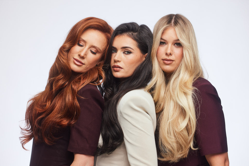 Three women posing together against a light gray studio background, showcasing three different hair colors: red, black, and blonde. They are wearing matching dark plum-colored short-sleeve tops.