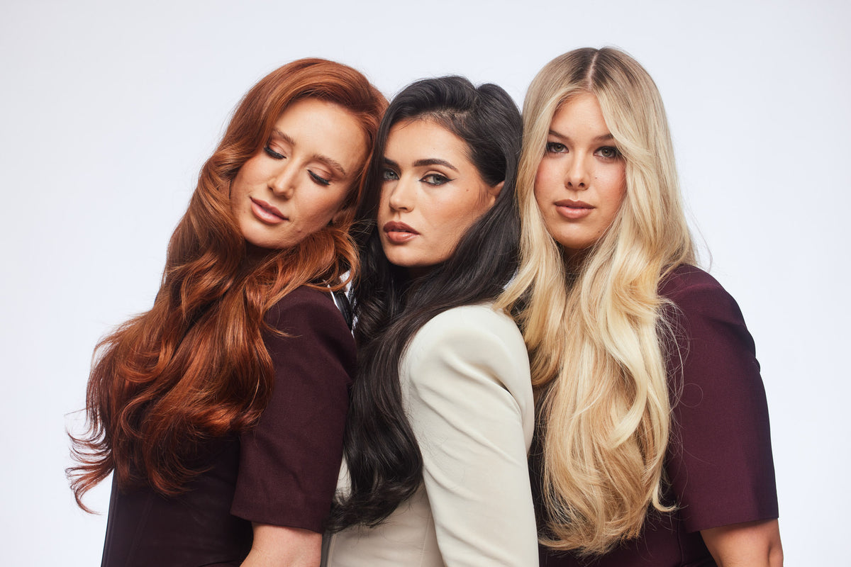Three women posing together against a light gray studio background, showcasing three different hair colors: red, black, and blonde. They are wearing matching dark plum-colored short-sleeve tops.