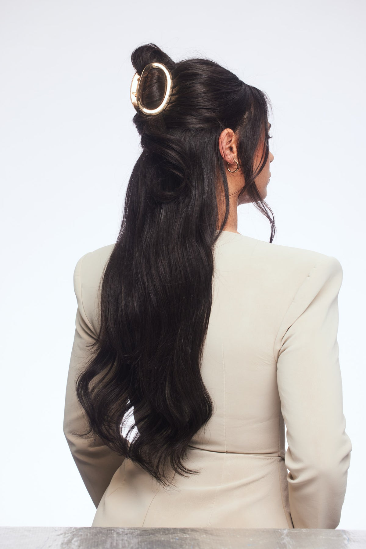 A woman shown from the back wearing a cream-colored blazer, with her long, dark wavy hair styled in a half-up bun secured by a gold ring hair accessory. She is photographed against a clean, light gray studio background.